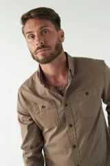 Camisa de hombre color beige con efecto stone wash, corte regular, cuello clásico, cierre frontal con botones, dos bolsillos con solapa abotonada en el pecho y mangas largas con puños abotonados.