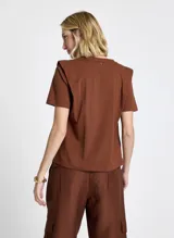 Remera de punto de algodón color terracota, de corte recto, largo estándar y mangas cortas. El diseño incluye superposiciones de tela en los hombros, creando un efecto estructurado y ligeramente geométrico. El cuello redondo presenta ribete acanalado y los laterales tienen pequeñas aberturas para un mejor ajuste.