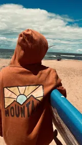 Hoodie color terracota/óxido con capucha y estampado gráfico en la espalda que representa un paisaje montañoso con un sol naciente o poniente y la palabra "MOUNTAIN" en la parte inferior.