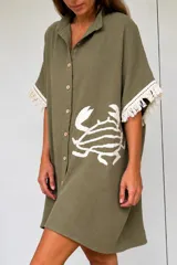 Túnica camisera corta de color verde oliva, de calce suelto y mangas cortas con flecos blancos. Cuenta con cierre frontal de botones y un diseño bordado de un cangrejo en la parte inferior.