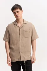 Camisa de manga corta color beige confeccionada en muselina de algodón, con cuello abierto, cierre frontal de botones y un bolsillo aplicado en el pecho.