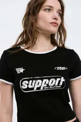 Remera baby crop de color negro con ribetes blancos en el cuello y las mangas. Cuenta con estampado frontal con texto "support your local team", estrella y el número "2001".