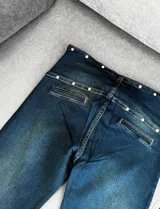 Pantalón jean de corte recto y tiro alto, color azul oscuro, con pretina ancha decorada con múltiples ojales metálicos y tachas, e incluye un cinturón incorporado con hebilla metálica.