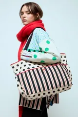 Bolso grande de tela con diseño combinado de lunares negros sobre fondo crema y bolsillo frontal con estampado de rayas verticales negras y rojas. Cuenta con ribetes en color rojo y correas negras.