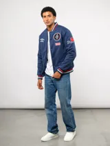 Campera bomber azul marino estilo retro, con cierre frontal de botones a presión, cuello, puños y ruedo con tejido acanalado tricolor. Presenta el logo de Umbro y el escudo del Club Nacional de Football bordados en el pecho, junto con el año 1899 y parches decorativos en las mangas.