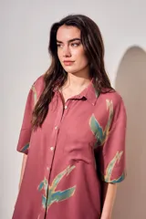 Vestido camisero corto de color rosado con estampado de aves abstractas en tonos verdes y azules. Cuenta con cuello camisero, cierre frontal con botones, mangas cortas y un bolsillo aplicado en el pecho.