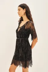 Vestido corto negro con mangas cortas de encaje, bordado a mano con pequeñas cuentas o lentejuelas. Presenta un escote pronunciado en forma de U con un corte de gota central y un cinturón fino bordado a juego. El ruedo es de encaje con terminación festoneada.
