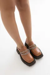 Mocasín de cuero color suela con punta cuadrada y suela track de goma oscura. Presenta un detalle de doble cadena metálica plateada sobre el empeine.