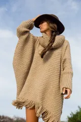 Poncho de tejido de punto grueso en color beige, con cuello alto tipo polera, mangas anchas y terminación con flecos en el ruedo.