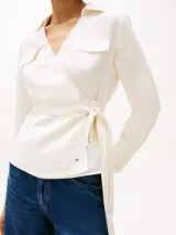 Blusa blanca de lino con diseño cruzado, cuello camisero, mangas largas con puños abotonados, dos bolsillos con solapa en el pecho y cinturón anudable.