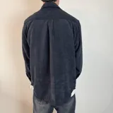 Sobrecamisa de pana negra con cierre frontal de botones, cuello clásico, mangas largas con puños abotonados y un bolsillo en el pecho.