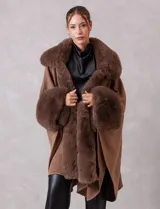 Ruana de tejido color tostado con cuello, bordes frontales y puños amplios con apliques de piel sintética al tono. Presenta una abertura central con cierre de botón tipo trenca.