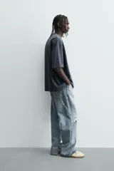 Camiseta de algodón color gris oscuro, corte relajado (relaxed fit), con cuello redondo y manga corta. Presenta dos inscripciones bordadas en hilo blanco en el frente.