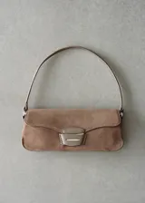 Bolso de hombro tipo baguette confeccionado en piel de serraje color tostado. Presenta un diseño rectangular con solapa frontal decorada con un detalle metálico y pespuntes visibles. Incluye un asa fija y forro interior.