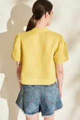 Blusa de lino color amarillo, con cuello mao, cierre frontal con cremallera y mangas cortas abullonadas. Cuenta con un bolsillo aplicado en el frente.