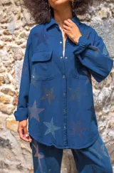 Camisa de algodón azul marino, corte oversize, con cuello camisero, manga larga y dos bolsillos de parche con solapa en el pecho. Toda la prenda está decorada con estrellas de pedrería.