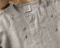 Blusa de lino color gris claro con bordados de pequeñas ramas en tono ocre. Presenta cuello redondo, cartera oculta con botones de nácar y mangas largas con puños estructurados con tablas.