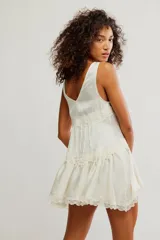 Mini vestido tipo slip de tela satinada negra, con escote en V profundo, diseño en capas con volados y detalles de encaje en los bordes. Corte estilo babydoll, sin mangas y silueta amplia.