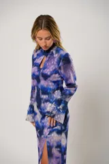Vestido largo con estampado abstracto en tonos azul, lila y blanco. Tiene cuello mao con abertura en forma de lágrima, mangas largas acampanadas, espalda abierta, tajo frontal y flecos en el ruedo.