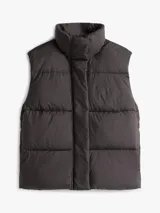 Chaleco acolchado de corte regular, confeccionado en poliéster reciclado. Presenta cuello alto con cierre de botones a presión, cierre frontal de cremallera oculto y diseño sin mangas. Incluye detalles distintivos y logo de la marca.