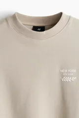 Buzo color beige de corte holgado, con cuello redondo y puños y ruedo acanalados. Estampado pequeño con la frase "New York" y un diseño de hojas en el pecho.