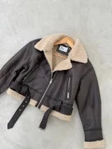 Campera tipo gamulán de cuero sintético en color marrón oscuro, con interior y detalles en corderito beige. Presenta cierre frontal, bolsillos laterales con cierre y cinturón ajustable en la cintura.