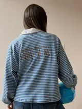 Buzo tipo chomba de corte oversize con diseño a rayas horizontales celestes y blancas. Presenta cuello camisero blanco y un bordado en la espalda con la inscripción República Oriental del Uruguay.