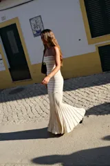 Vestido largo de punto color beige, sin tirantes y con escote palabra de honor. Presenta un diseño ajustado en el torso y falda amplia de corte sirena con pliegues acampanados en la parte inferior.