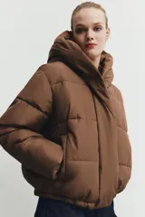 Campera puffer acolchada de color negro, con cuello envolvente, capucha integrada, manga larga, bolsillos laterales ocultos y bajo con elástico. Cierre frontal con botones a presión ocultos por solapa.
