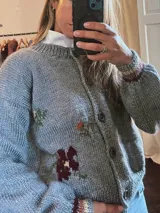 Sweater de punto gris con diseño de flores bordadas, cuello redondo y cierre frontal con botones. Presenta puños con detalles de rayas en tonos tierra.
