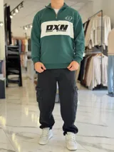 Hoodie de corte clásico con medio cierre y cuello alto, en color marrón con una franja horizontal blanca en el pecho que incluye el logo estampado en negro. Presenta además un pequeño bordado en el hombro izquierdo y puños y cintura elastizados de rib.