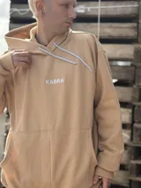 Canguro de tela deportiva color beige, con capucha, cordones ajustables en color blanco y logo bordado en el pecho.