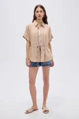 Camisa de viscosa color beige, con cuello camisero, mangas cortas, cierre frontal con botones ocultos y cordón ajustable en la cintura.