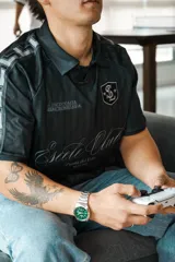 Camiseta de fútbol estilo boxy de manga corta y cuello polo, en color negro con sutiles rayas verticales. Presenta inscripciones estampadas y escudo bordado en el pecho.