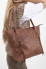 Cartera tipo tote bag Sagali, confeccionada en 100% cuero vacuno en tono marrón con efecto gastado. Presenta doble asa de hombro, detalles de tientos trenzados en los laterales con flecos colgantes, costuras verticales decorativas y placa metálica con el logo de la marca en el frente.