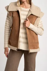 Chaleco estilo gamulán de corte oversize, confeccionado en material tipo gamuza color camel con interior y bordes de corderito sintético color crema. Presenta cierre frontal completo, bolsillos laterales y detalles de tiras decorativas en los hombros.