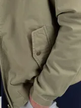 Campera Harrington color beige, con cuello alto abotonado, cierre frontal completo y bolsillos laterales con solapa y botones a presión. Puños y cintura elastizados de rib.