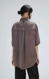 Camisa de corte oversize y mangas cortas, confeccionada en tela con efecto de teñido artesanal maltinto en tono gris. Presenta cuello clásico, cierre frontal con botones y costuras verticales decorativas en el frente.