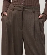 Pantalón de vestir estilo sastre con corte wide leg, confeccionado en tejido crepé color marrón con sutil estampado de rayas verticales. Presenta pliegues delanteros, cintura estructurada con trabillas y cierre frontal.
