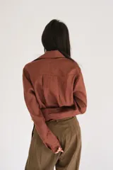 Camisa blanca de manga larga, con botones de guampa y un detalle de roseta o flor a tono en la parte delantera. El calce es holgado con la espalda ligeramente más larga.