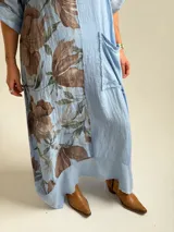 Vestido largo celeste con gran estampado floral marrón en el lateral derecho, de corte holgado, mangas 3/4 con trabilla y dobladillo irregular con flecos desflecados. Incluye un bolsillo aplicado en el frente.