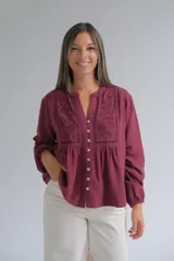 Blusa de manga larga color bordó con cuello mao y cierre frontal de botones. Presenta detalles de bordados florales y calados en el frente, con un corte holgado y puños elásticos.