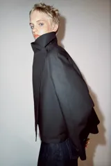 Cazadora corta de color negro con cuello subido cruzado, manga larga con puños anchos ajustados con trabilla y botón, y cierre frontal cruzado con botones ocultos.