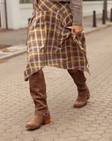 Botas de caña alta confeccionadas en cuero gamuzado color marrón. Presentan un diseño de estilo western con puntera cuadrada, taco de madera y una tira decorativa con estribo metálico desmontable en la zona del tobillo.
