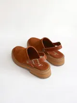 Zueco de cuero gamuzado color óxido, con tira trasera ajustable mediante hebilla metálica. Presenta una suela de plataforma con textura acanalada y detalle de textura reptil en la tira posterior.