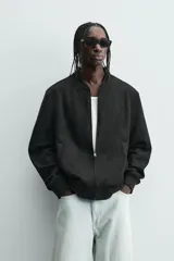 Cazadora bomber negra de corte regular fit, confeccionada en tejido efecto ante. Cuenta con cuello, puños y bajo en rib elástico, manga larga, bolsillos de vivo en la cadera y un bolsillo interior. Cierre frontal con cremallera.