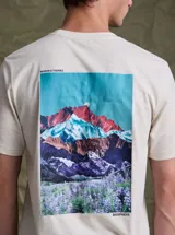 Remera de algodón color beige jaspeado, con estampa fotográfica en la espalda que muestra un paisaje de montañas coloridas y campo de flores.