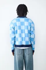 Sweater de tejido de punto con diseño a cuadros en tonos celestes y azules. Presenta cuello en V con solapa, mangas largas y detalles estampados al frente con estrellas y texto deportivo.