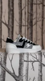 Championes urbanos con diseño de rayo en el lateral, combinando paneles de glitter plateado con detalles en gamuza negra y cuero sintético blanco. Poseen cordones blancos y suela de goma blanca.