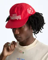 Gorra de béisbol de cinco paneles sin estructura color rojo, con visera plana y bordado frontal en tono crudo con la frase 'I NEED Therapy'. Presenta detalles bordados adicionales y correa ajustable de nailon en la parte trasera.